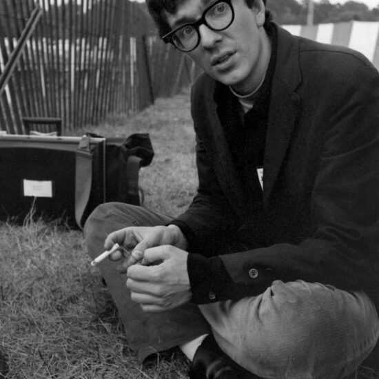 Photographer Adam Ritchie sits cross-legged on the grass and holds a cigarette, backstage during the Newport Jazz Festival, Rhode Island, July 1965