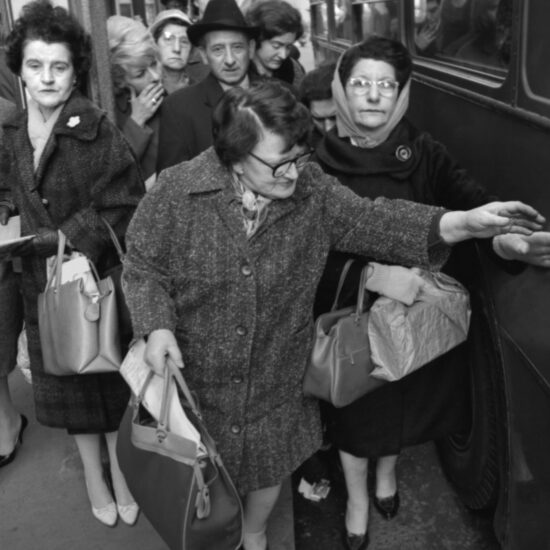 A queue of people in winter clothes start boarding a bus at a stop outside Aldgate East underground station that serves routes 10, 25 and 32 heading to Whitechapel, Mile End, Bow and Stratford. Women carry large handbags: several people stare at the camera with a weary look; London, circa Spring 1964