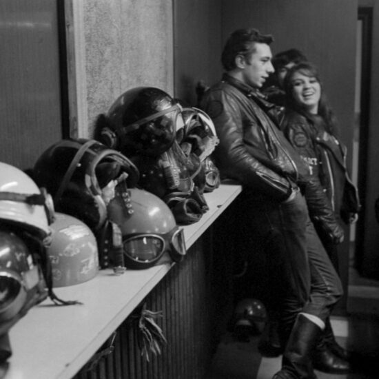 Male and female bikers laugh with each other by a shelf covered in motorbike helmets in the Ace Cafe, circa 1964