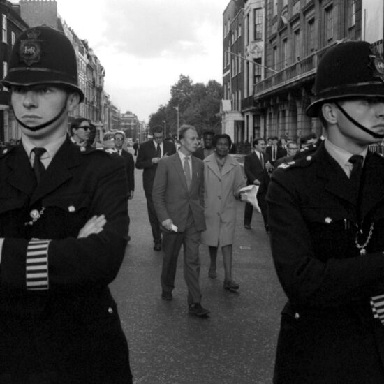 Two white police officers stand in the foreground as 'Black Equality' march leaders and demonstrators walk through Mayfair streets to the American Embassy, central London, circa 1963