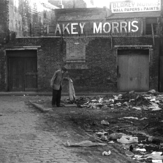 An elderly man sorts through rubbish in front of the Blakey Morris Wallpapers and Paints factory in south London, circa 1960
