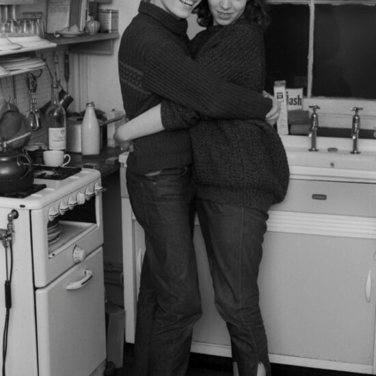 A  young white man and woman dressed in beatnik/bohemian clothes, hug each other in front of a sink in a typical 1960's British kitchen. She is barefoot and they both wear denim jeans and knitted jumpers