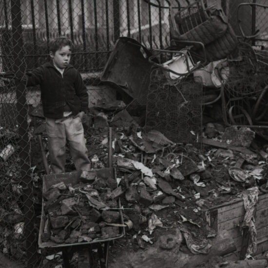 A small boy stands on top of a pile of rubble and broken furniture, collecting items in a wheelbarrow during construction of an adventure playground on derelict land in Notting Hill, West London, circa 1962