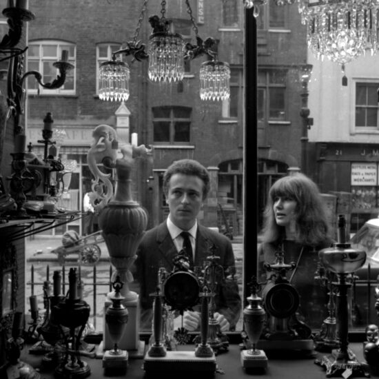 A man and a woman look at antiques in a shop window, photographed from inside the premises. There are many ornate antique items in the shop front including sconces, candelabra, clocks and small glass electric chandeliers. There is a handcart visible in the street behind the couple, and several ornate buildings with shops on the ground floor; London circa 1964