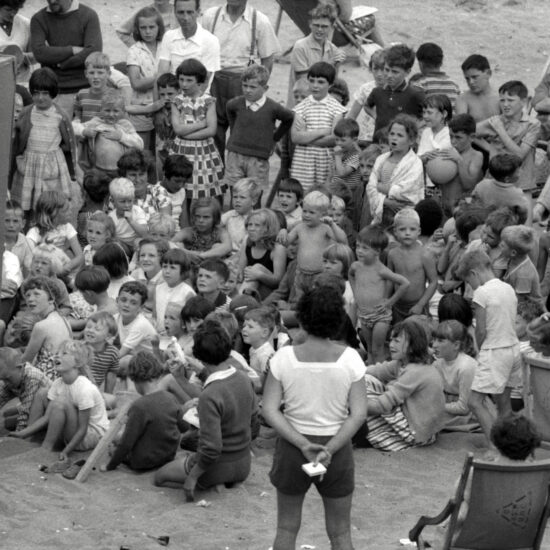 During a CND outing to the beach, children and adults gather around a Punch and Judy tent on the sand. The children are transfixed by the off-screen puppets, circa 1963