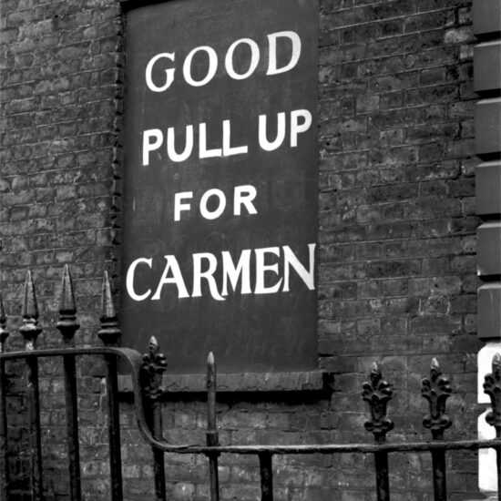 Behind broken railings a sign on a boarded up window reads Good Pull Up For Carmen, London circa 1961