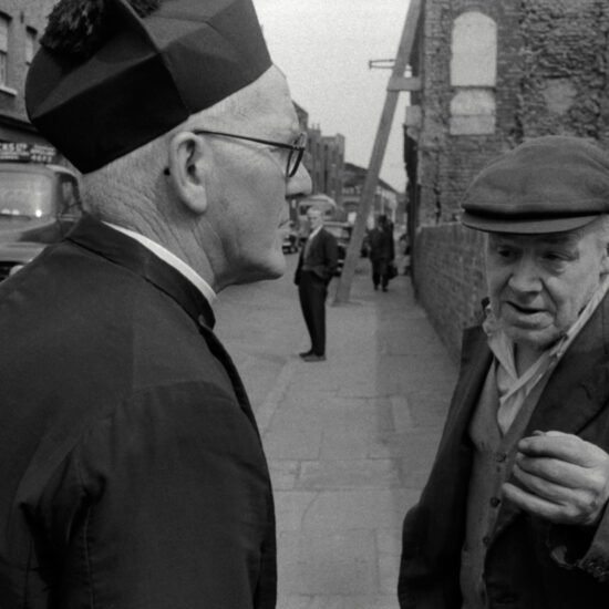 A Catholic priest wearing a black biretta talks to an elderly man wearing a flat cap in a bombed-out street in the East End of London: in the background is a GB Hicks Ltd. lorry