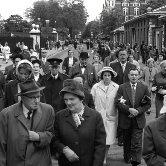 A crowd of smartly dressed people walk down the street towards the photographer as they near the entrance to the 1962 Chelsea Flower Show