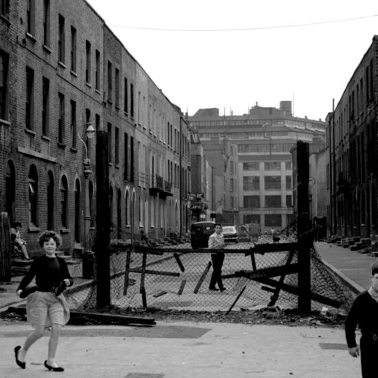 A group of children play in a London street which still has a blockade left over from the Second World War; one girl is wearing her mother’s fancy shoes, 10 April 1961