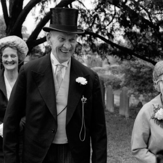 A male guest in top hat and tails accompanies two female guests wearing hats as they arrive at the church for a country wedding