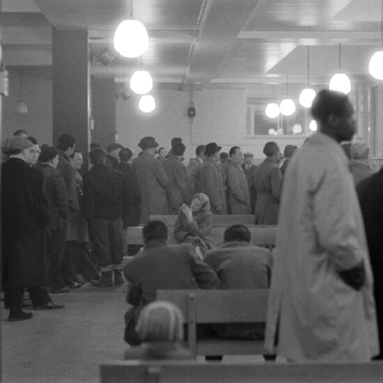 A woman on a bench is surrounded by men queueing at a Men's Unemployment Benefits Office counter, London, circa 1963