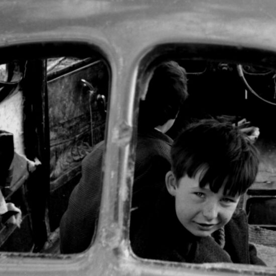 Two young boys play in an abandoned and gutted motor car on a demolition site in London’s East End. One boy stares out of the missing rear window at the viewer: in the background boarded up and empty terraces of WWII bomb-damaged houses sit beside new low-rise blocks of flats; London, circa 1964