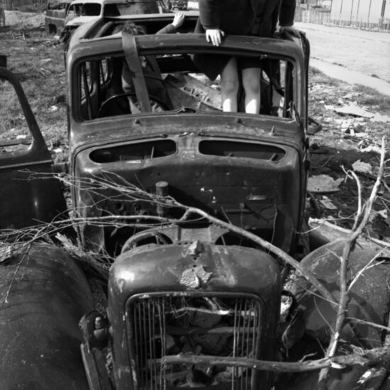 Two young boys play in an abandoned and gutted motor car on a demolition site in London’s East End. One boy is climbing out of the missing roof panel: in the background other stripped vehicles lay abandoned in front of terraces of WWII bomb-damaged houses; London, circa 1964