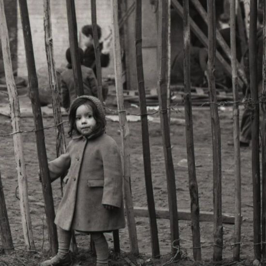 A small girl in a thick overcoat stands in front of a tall fence on derelict land in Notting Hill, West London, circa 1962