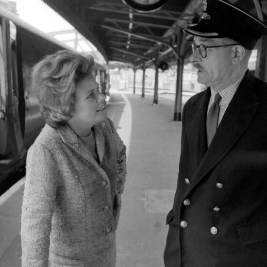 A blonde female talks to the Station Master on the platform at Paddington Railway Station. She has one hand on a bundled stack of newspapers and smiles at the Station Master - there is a train behind them with an open door