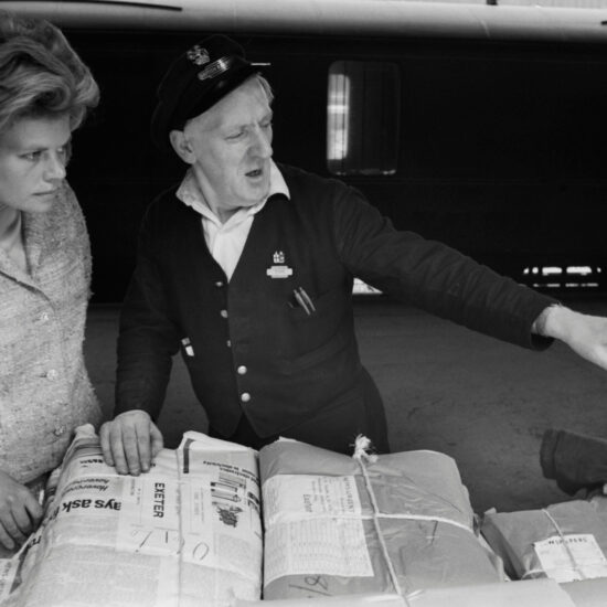 A blonde female talks to a railway worker on the platform at Paddington Railway Station as he points at something off camera. They are standing in front of a train and behind bundled stacks of The Times newspapers, destined for Exeter