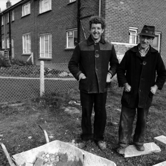 Two male labourers in donkey jackets pose by their wheelbarrow on a post-war housing estate in Frimley and Camberley, Surrey, circa 1963