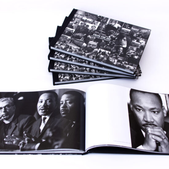 Five copies of the book FROM THE HIP - Photographs by JOHN 'HOPPY' HOPKINS 1960-66 on a white background. Four copies are stacked in a pile, and an open copy of the book in the foreground features images of Canon Collins, Dr Martin Luther King and Reverend Ralph Abernathy on one page and a close-up of Dr Martin Luther King on the other.
