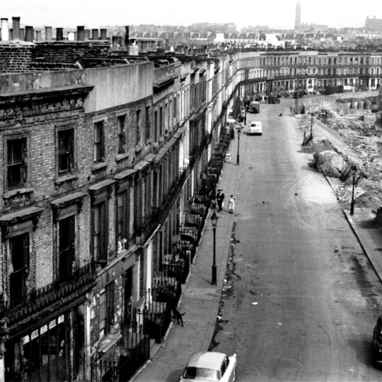 An aerial view of Harrow Road, including tenement buildings, an undeveloped bomb-site and several churches in the background, London 1961