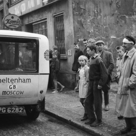 The yellow hearse is parked outside a police station in Poland. Onlookers stare at the vehicle and at the photographer. Circa 1960