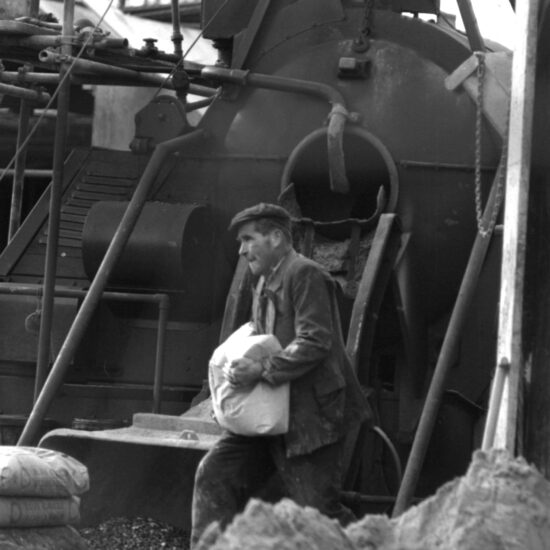 A man in a cloth cap and tattered suit carries a heavy sack in front of scaffolding and weighing machinery on a building site, England circa 1963