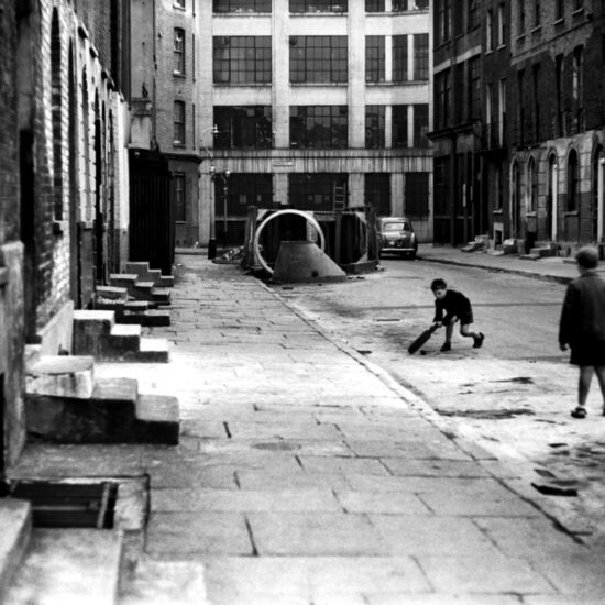 Two boys in shorts play cricket in the road, with working-class terraced housing on both sides and an industrial building sited on Ebenezer Street in the background, East London circa 1961