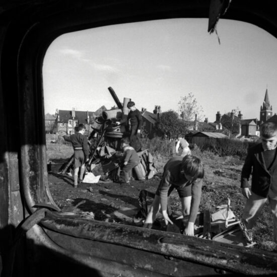 Framed by a door window, a gang of small boys play near an abandoned car and build a bonfire on a patch of waste ground in Kingston, West London, circa 1961