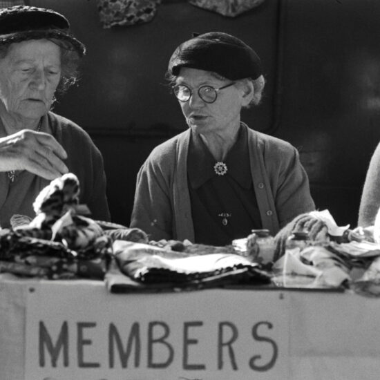 Three older women wearing hats sit behind a table marked 'Members' that carries knitted items in Kingston, west London, circa 1961