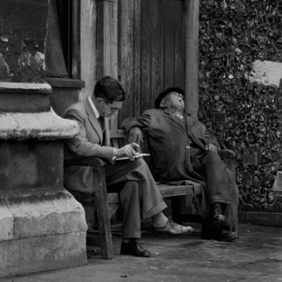 An elderly man snoozes whilst a younger man reads on a bench in Kingston, west London, circa 1961