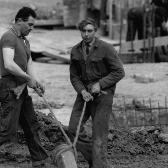 Two labourers working on a building site carry a long metal strut with a rope: one wears a boiler suit and the other is in jeans and a polo shirt. There are other labourers working behind vertical rebars in the background, London circa 1963