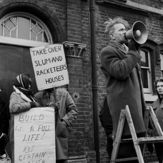 A man stands on a step-ladder and addresses a crowd through a megaphone, during a housing demonstration against racketeer landlords in Westmoreland Road, Southwark, London, in the freezing winter of 1963. One demonstrator holds a sign saying 'Take Over Slum And Racketeer Houses'