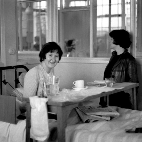 Ann James smiles and sits up in bed in a maternity ward in a London NHS hospital. She has a tray with crockery and newspapers on her bed. A woman standing by the bed looks away from camera, circa 1960/61.