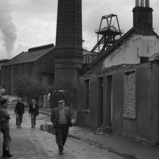 Men walk along a street past a derelict building with a colliery wheel in the background, in the mining village of Kinglassie, Fife, Scotland, circa 1963