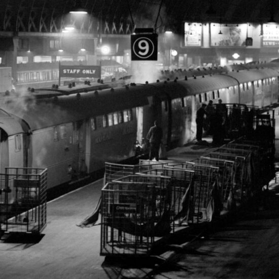In 1966 men load the Paddington mail train at night, a wide shot from overhead including advertisements for The News of the World in the background