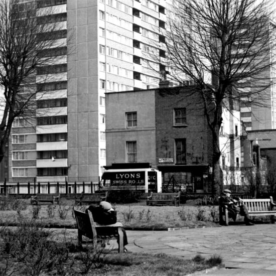 Two older men rest on benches in an open space with Victorian terraced buildings, new-build tower blocks of flats and a Lyons Swiss Rolls delivery van in the background, London 1961