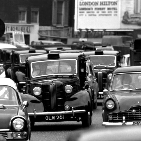 Old fashioned black London taxi-cans and motor cars are stuck in a traffic jam on Park Lane: in the distance there is a constructor’s hoarding for the London Hilton Hotel; London circa 1962
