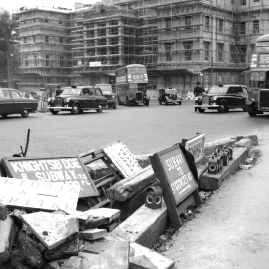 A pile of obsolete wooden directional signs for various central London locations lie on the ground in a pile at the top of Park Lane on Hyde Park Corner. In the distance a police officer directs traffic flow: London circa 1962