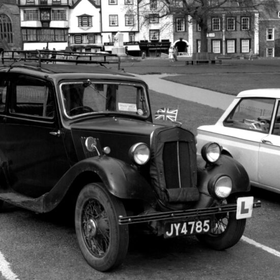 A black vintage motorcar sits in a parking space on Parliament Square: it has a Union Jack British flag stuck into the front grille and a learner driver L plate tied to the front bumper. London circa early 1964