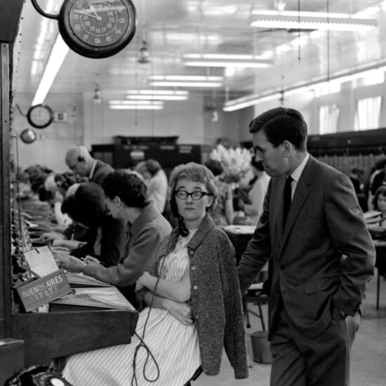 In 1964 a male supervisor chats with the female telephone exchange operator responsible for Buenos Aires connections as she sits at her desk. There are many exchange operators working in the background of the shot