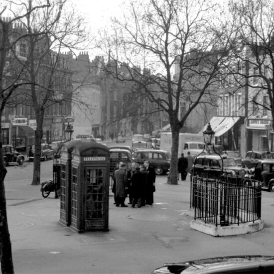 A group of men stand and chat by several public telephone boxes on Pimlico Road, with stores and the offices of the Grosvenor Estate in the background, London circa 1960/61