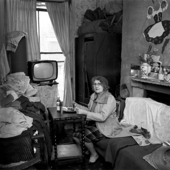 A middle-aged woman in a cramped bedsit wears her coat to keep warm as she eats her dinner at a small table in between her bed and the sofa, with a television in front of the window, Harrow Road, London circa 1964