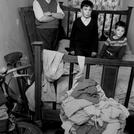 A man stands and two boys sit on the bed in a tiny room, with a bicycle and a pile of clothes in the foreground. Harrow Road, London, circa 1964