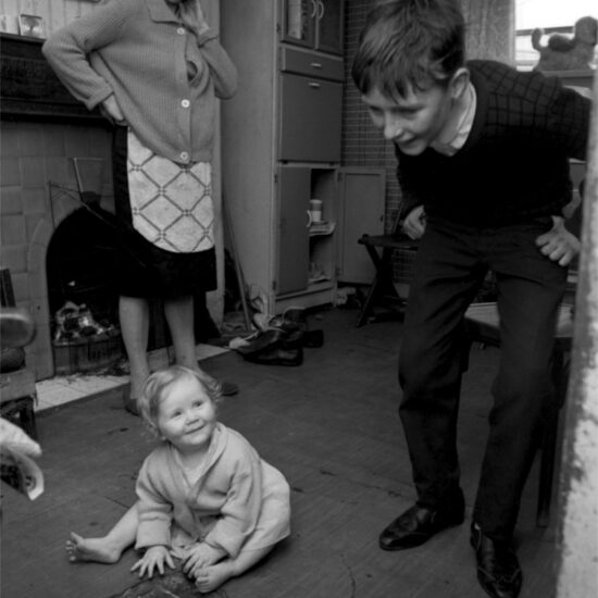 A smiling toddler sits on the floor and pulls at damaged linoleum whilst a boy looks on and a woman stands in front of a coal fire. Harrow Road, London circa 1964