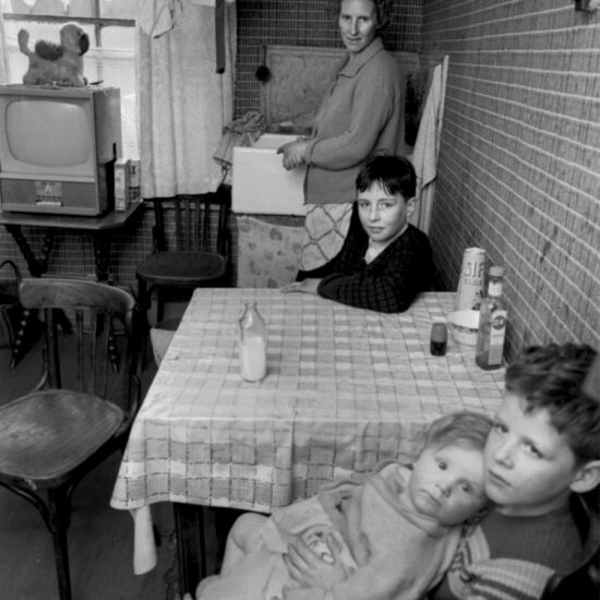 A woman stands at a sink and two boys sit at a kitchen table and one boy holds a toddler. Harrow Road, London circa 1964