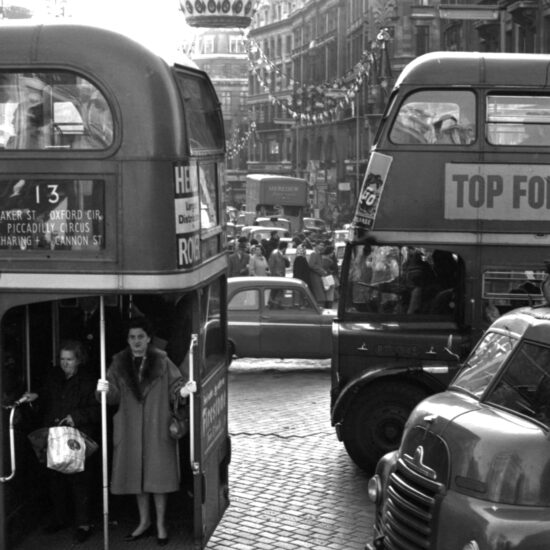 Two women wait to disembark an open-platform number 13 Routemaster double-decker bus as it travels south along Regent Street, past pedestrians, Christmas festive street decorations, a Meredew Furniture van, motor cars and taxi-cabs; London, December 1961