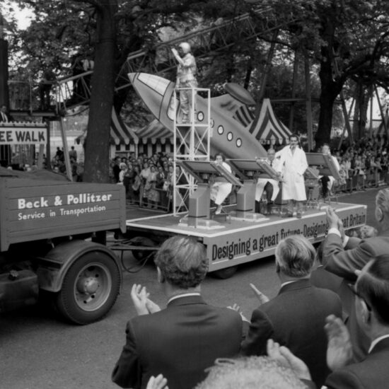 During the 1962 Festival of Labour in Battersea Park, a float takes part in a parade. The float is titled 'Designing a great future - Designing a Great Britain' and has people dressed as scientists and astronauts working at draughting tables, as well as a futuristically-styled rocket ship. The float is passing Labour Party leaders, seen from the back