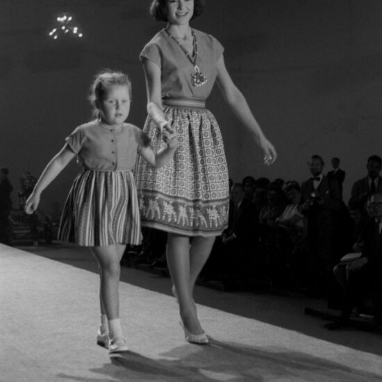 A woman and a girl walk down a catwalk in front of photographers during a fashion show at the USSR Industrial Exhibition in London, held at Earl's Court, 07-29 July, 1961