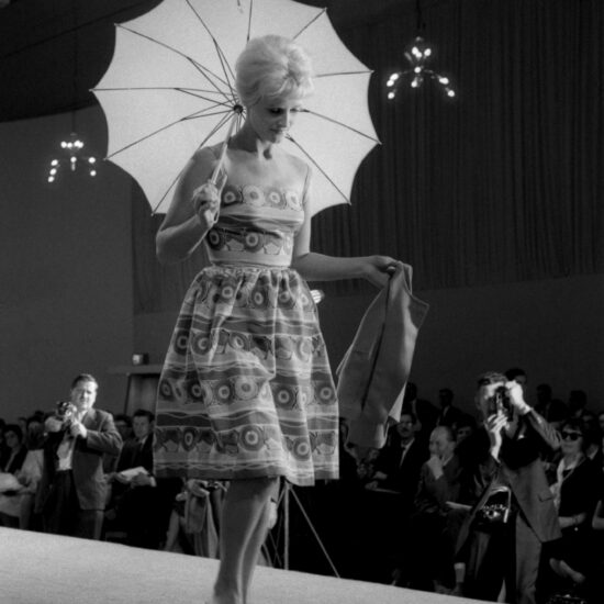 A model carrying an open umbrella walks down a catwalk in front of photographers during a fashion show at the USSR Industrial Exhibition in London, held at Earl's Court, 07-29 July, 1961