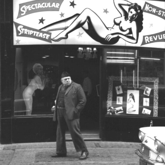 A man walks past a striptease joint, looking behind him with a grumpy expression. There are photographs of the performers in seductive poses in the window, below an illuminated sign which reads ‘Spectacular Striptease Non-stop Revue’, Soho, London, circa 1964