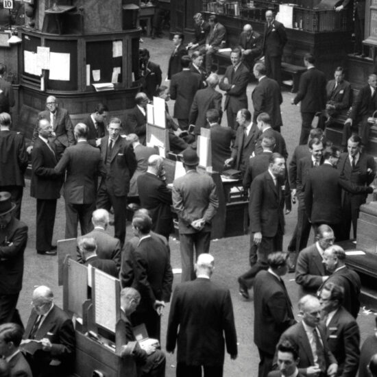 An overhead shot of the floor of the Stock Exchange, featuring wood panelling, men conversing, trades being made and noticeboards displaying the date (May 24 1961)